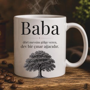 以下が含まれることがあります： 白いセラミックコーヒーマグ。大きな黒い文字で「Baba」と書かれています。その下にテキストと白黒の木のイラストがあります。マグカップから湯気が立ち上り、温かい飲み物を連想させます。