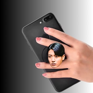 Peut inclure: Un accessoire de smartphone noir doté d'un dessus circulaire avec le portrait imprimé d'une personne aux cheveux foncés. L'accessoire est fixé au dos d'un smartphone noir, offrant une prise pour maintenir l'appareil fermement en main.