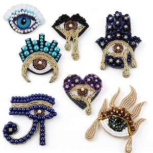 Puede incluir: Colección de broches con forma de ojo adornados con cuentas en varios diseños. Los broches presentan intrincados trabajos de cuentas en colores como azul, dorado, negro y morado. Algunos diseños incluyen detalles de perlas y lágrimas.