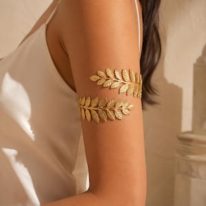 May include: A gold-toned metal arm cuff featuring a wrap-around design with two distinct leaf-shaped branches. The accessory is worn on the upper arm, showcasing a textured, botanical-inspired pattern that encircles the limb in a spiral shape.