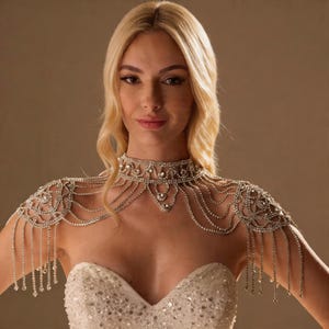 May include: A dazzling silver shoulder necklace, adorned with numerous rhinestones. The design includes a choker-style collar and flowing strands that cascade over the shoulders. The necklace has a glamorous, elegant appearance, with delicate fringe detailing.