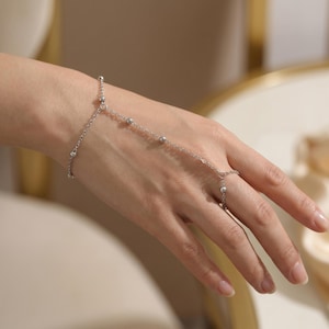 Puede incluir: Una delicada pulsera de cadena de mano plateada. La pulsera presenta pequeñas cuentas redondas conectadas por finas cadenas. Una cadena se extiende hasta un anillo, mientras que otra se conecta a la muñeca. La joya se muestra en una mano.