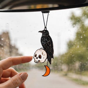 May include: A hanging car ornament featuring a black raven perched atop a white human skull, with a small orange crescent moon dangling from the bottom. The piece is constructed with dark metal outlines and translucent colored sections.