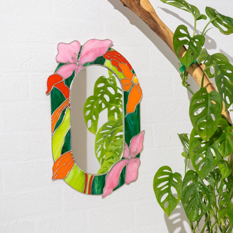 May include: An oval wall mirror featuring a stained glass frame decorated with vibrant pink, orange, and lime green floral and leaf motifs. The glass pieces are joined by thin metal soldering, creating a colorful border around the central reflective surface.