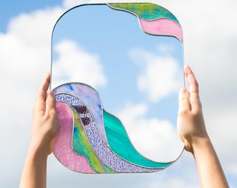 May include: A rectangular mirror with rounded corners featuring decorative stained glass accents in pink, green, blue, and purple along the top right and bottom left edges, framed with thin metal solder lines.