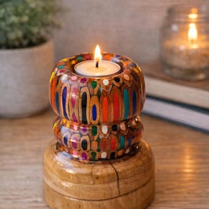 Può includere: Un portacandele artigianale in legno, con un design unico. La sezione superiore è decorata con pezzi di matite colorate, creando un effetto mosaico. Una candela da tè accesa si trova al centro, emettendo una luce calda.