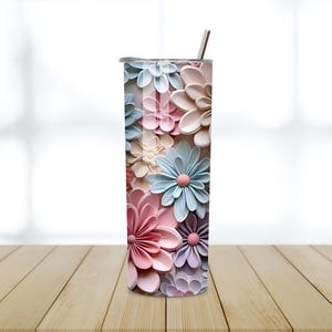 Puede incluir: Un vaso alto de acero inoxidable con un dise&ntilde;o floral. Las flores son en tonos de rosa, azul y crema, con un efecto 3D. El vaso tiene una pajita plateada y una tapa. El fondo est&aacute; borroso.