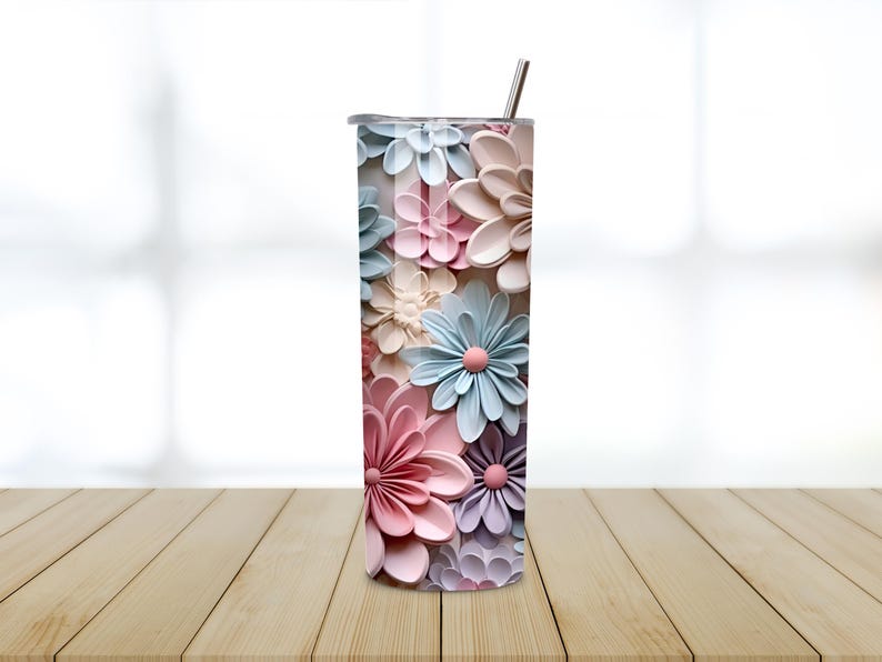 Puede incluir: Un vaso alto de acero inoxidable con un dise&ntilde;o floral. Las flores son en tonos de rosa, azul y crema, con un efecto 3D. El vaso tiene una pajita plateada y una tapa. El fondo est&aacute; borroso.