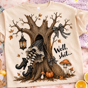 May include: A beige t-shirt featuring a graphic of a tree with a witch's legs protruding from a hollow, wearing black and white striped stockings and buckled boots. A lantern, a broom, mushrooms, and the text "Well, shit..." are printed on the front.