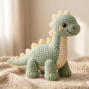 May include: A handmade crochet dinosaur toy in green and cream. The dinosaur has a smiling face and yellow spikes along its back. The toy is made with a textured stitch, and the belly is a contrasting cream colour.