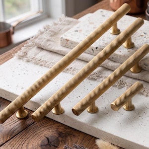 Può includere: Una collezione di maniglie per mobili color oro con un motivo a diamante strutturato. Le maniglie sono di varie lunghezze, con un design cilindrico e basi arrotondate. Sono esposte su una superficie bianca maculata, suggerendo un tema di ferramenta per cucina o mobili.