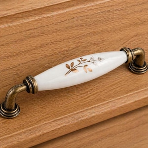 May include: A white ceramic cabinet pull with a floral design in gold and brown tones. The handle has a curved shape with antique brass-colored metal ends. The pull is mounted on a wooden surface.