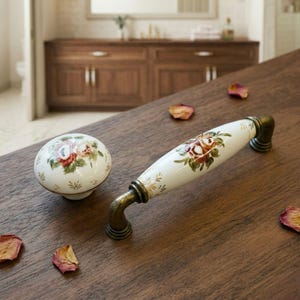 May include: A white ceramic cabinet knob and pull with floral designs in red, pink, and green. The hardware has antique brass-colored metal accents. The knob is round, and the pull is a long, curved handle.