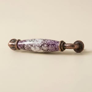 May include: A cabinet pull with a marbled purple and white center and bronze-colored metal ends. The pull has a cylindrical shape with decorative details on the ends. This hardware is suitable for drawers and cabinets.