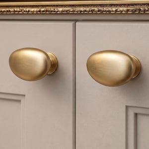 Puede incluir: Primer plano de dos pomos de armario ovalados de latón cepillado montados en una puerta de armario gris claro. Los pomos tienen un diseño suave y redondeado y un acabado metálico cálido. La puerta del armario tiene un diseño de panel empotrado.