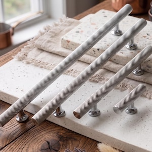 Puede incluir: Un conjunto de cinco tiradores plateados para armarios con un patrón de diamantes texturizado. Los tiradores se muestran sobre una superficie blanca moteada, resaltando su diseño. Adecuados para muebles y decoración del hogar.