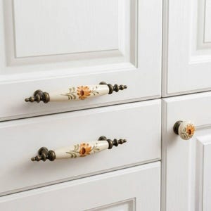 Puede incluir: Tiradores de armario color crema y un pomo con diseño floral. Los tiradores tienen una base y tapas de color bronce, con un centro color crema con un estampado floral naranja y verde. El pomo tiene un diseño similar.