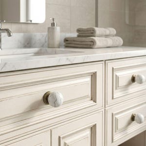 May include: Cream-colored bathroom vanity with marble-look knobs and a marble countertop. The vanity has four drawers with decorative molding. A chrome faucet, soap dispenser, and folded towels are visible.