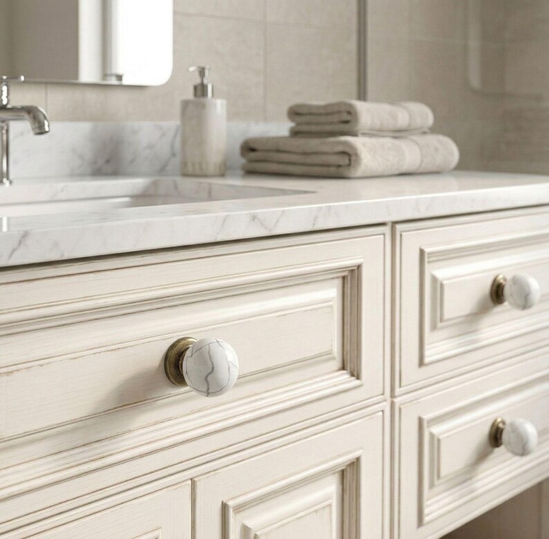 May include: Cream-colored bathroom vanity with marble-look knobs and a marble countertop. The vanity has four drawers with decorative molding. A chrome faucet, soap dispenser, and folded towels are visible.