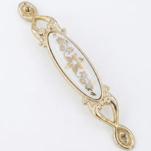 May include: A gold-toned cabinet pull with a white oval center. The center features a gold floral design. The pull has ornate detailing on each end and around the oval center. Hardware for cabinets and drawers.
