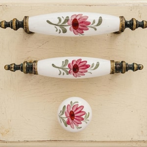 May include: White ceramic cabinet pulls and knob with a floral design. The pulls are oval-shaped with bronze-coloured metal ends and gold accents. The floral design features a red flower with green leaves. The knob is round with the same floral design.