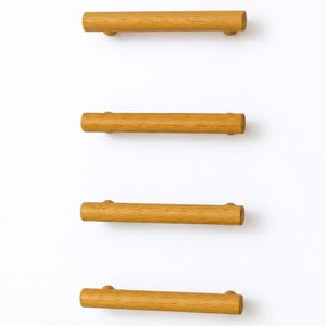 May include: Four wooden cabinet pulls with a light brown finish are arranged vertically against a white background. Each pull is cylindrical with rounded ends, designed for easy gripping and opening of drawers or cabinets.