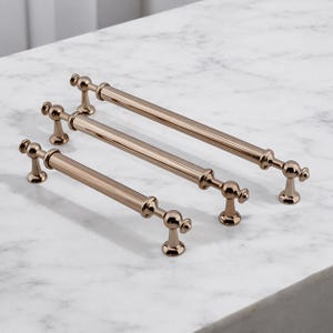 May include: Three bronze-colored cabinet pulls of varying lengths are arranged on a white marble surface. Each pull features a cylindrical bar with decorative end pieces and a central accent. The hardware is designed for furniture.