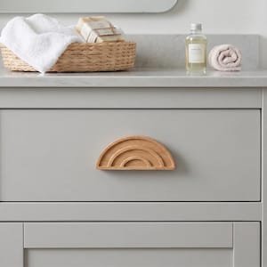 May include: A light gray bathroom cabinet drawer with a wooden rainbow-shaped handle. A woven basket with white towels and soap bars sits on the countertop. A bottle of bath oil and a rolled towel are also on the counter.