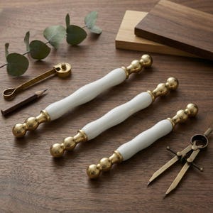 Può includere: Tre maniglie per mobili bianche e dorate con finitura in ottone sono disposte su una superficie di legno. Sono presenti anche un piccolo utensile in ottone, una matita e un compasso. Le maniglie hanno un centro bianco curvo e estremità sferiche dorate.