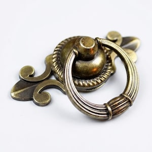 May include: Antique brass drawer pull with a ring handle. The decorative hardware features a detailed design with a circular handle and ornate backplate. The finish has a vintage look, suitable for cabinets, drawers, or furniture restoration projects.