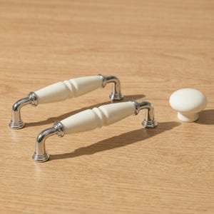 Puede incluir: Dos tiradores de armario color crema y un pomo a juego. Los tiradores tienen un diseño curvo y acanalado con detalles metálicos plateados pulidos. El pomo es redondo con una base corta y cilíndrica. Todo el conjunto está sobre una superficie de madera clara.