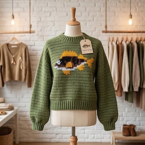 May include: An olive green crochet sweater with a crew neck and long sleeves, adorned with a fish motif in yellow, brown, and white. The sweater is displayed on a mannequin with a "Handcrafted" tag. Other garments are visible in the background.