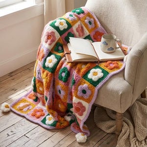 May include: A colorful, crocheted blanket with flower motifs in shades of pink, orange, green, and white is draped over a beige armchair. An open book and a mug with cloud designs rest on the blanket. The blanket has a lilac border and pom-pom accents.
