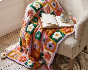 May include: A colorful, crocheted blanket with flower motifs in shades of pink, orange, green, and white is draped over a beige armchair. An open book and a mug with cloud designs rest on the blanket. The blanket has a lilac border and pom-pom accents.