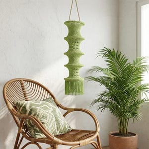 May include: A green, handmade macrame pendant light fixture with a tiered design and fringe detail. The light hangs from a natural rope and is suspended in front of a white wall. A rattan chair and potted plant are in the foreground.