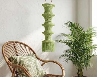 May include: A green, handmade macrame pendant light fixture with a tiered design and fringe detail. The light hangs from a natural rope and is suspended in front of a white wall. A rattan chair and potted plant are in the foreground.