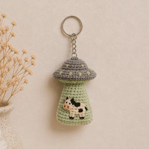 May include: A handmade crochet keychain featuring a gray and green UFO design with a small cow applique. The keychain has a silver metal ring and chain. The UFO has a gray top with green dots and a green base.