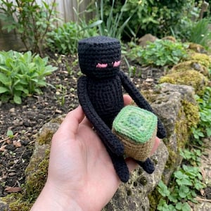 May include: A black crochet Enderman figure from the Minecraft game, with pink eyes and a green and brown block. The figure is held in a hand, with a garden in the background.