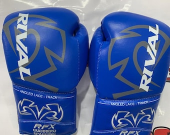 Op de afbeelding: Een paar blauwe bokshandschoenen met de witte tekst RIVAL en een grijs logo. Op de pols staat de tekst ANGLED LACE - TRACK en RFX GUERRERO SPARRING. De handschoenen hebben een gladde afwerking en zijn bedoeld voor sparren.