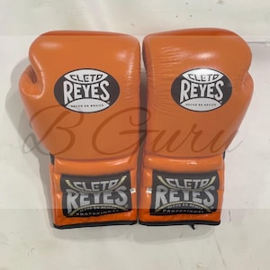 Könnte beinhalten: Ein Paar orangefarbene Boxhandschuhe. Jeder Handschuh trägt den Markennamen "CLETD REYES" in einem schwarzen Oval mit weißem Text. Die Handschuhe sind aus Leder und haben ein professionelles Design.