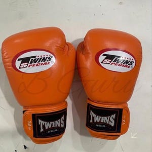 Könnte beinhalten: Ein Paar orangefarbene Boxhandschuhe mit dem Logo "Twins Special" in Weiß und Rot. Die Handschuhe haben schwarze Handgelenksriemen mit dem Logo "Twins Special" und Größenangaben von 14 oz und 12 oz.