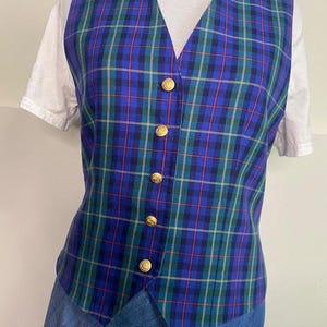 May include: A blue, green, and red plaid vest with gold buttons. The vest is worn over a white t-shirt and blue denim skirt. The vest has a V-neck and is buttoned up the front.