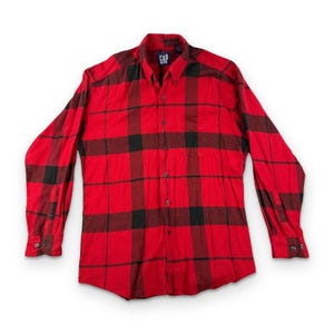 May include: A long-sleeve button-down shirt featuring a red and black plaid pattern. The shirt has a pointed collar, a front button placket, and buttoned cuffs. A blue Gap brand label is visible on the inside of the collar at the neckline.