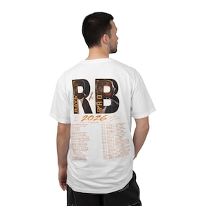 May include: A white short-sleeve t-shirt featuring a graphic on the back with the large letters RB, portraits of two individuals, the text RAYM & BRO, 2026 TOUR, and a list of tour dates and locations printed in orange and black ink.
