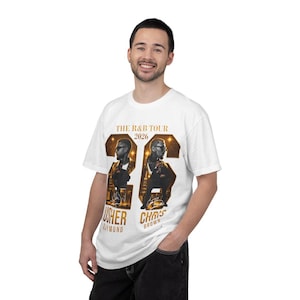 May include: A white short-sleeve t-shirt featuring a gold graphic print that reads THE R&B TOUR 2026. The design includes large gold numbers 26, images of Usher Raymond and Chris Brown, and their names printed in gold lettering below the figures.