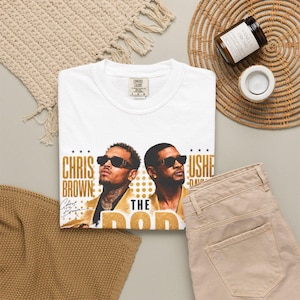 May include: A white short-sleeve t-shirt featuring a graphic print of Chris Brown and Usher wearing sunglasses. The text reads Chris Brown, Usher, and The Pop. The shirt has a Comfort Colors brand label at the inner neckline, indicating a size large.