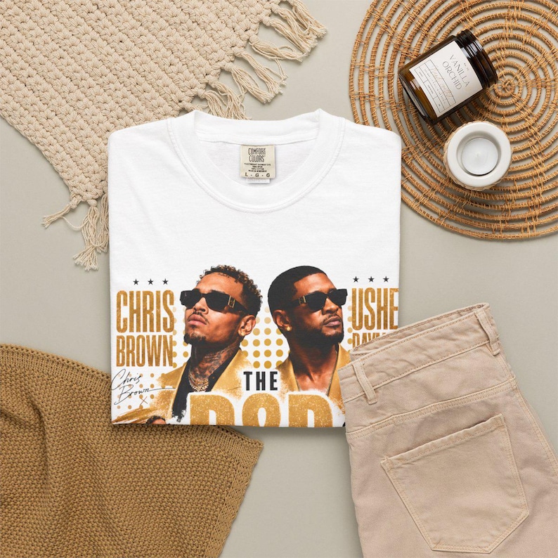 May include: A white short-sleeve t-shirt featuring a graphic print of Chris Brown and Usher wearing sunglasses. The text reads Chris Brown, Usher, and The Pop. The shirt has a Comfort Colors brand label at the inner neckline, indicating a size large.