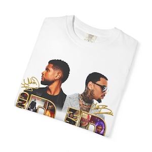 May include: A white t-shirt featuring a graphic print of two men, Usher and Chris Brown, in profile. The design includes gold cursive signatures, the text "USHER HERE | STAND", and smaller inset images of the artists in a collage-style layout.
