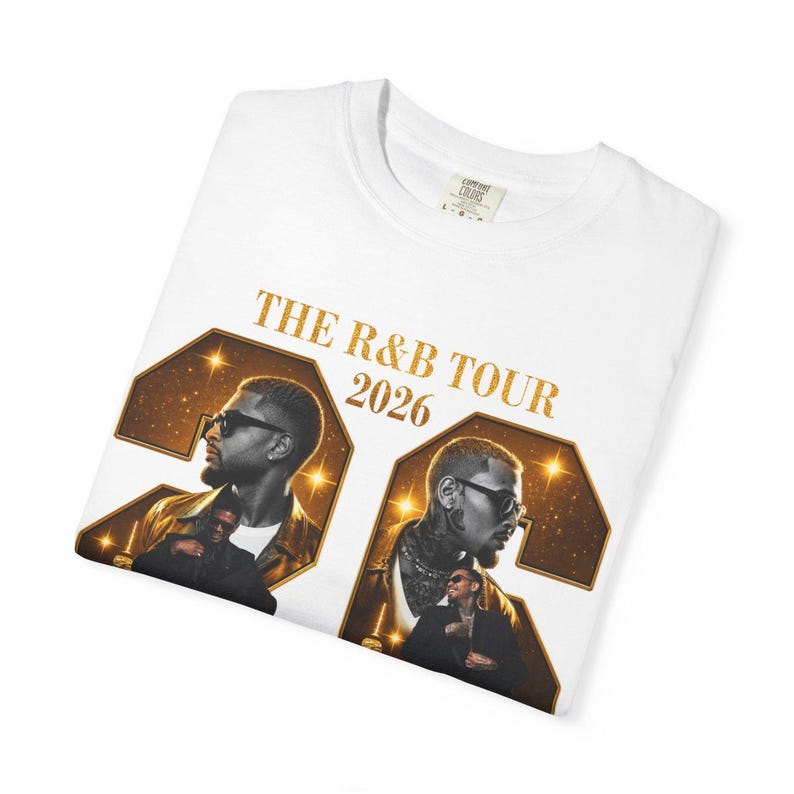 May include: A white t-shirt featuring a graphic print with the text THE R&B TOUR 2026. The design includes two large gold-outlined numbers containing portraits of two men wearing sunglasses, set against a dark, starry background with gold accents.