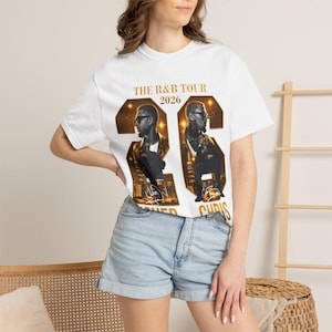 May include: A white short-sleeve t-shirt featuring a graphic print with the text THE R&B TOUR 2026. The design includes two large gold-toned numbers forming a 26, each containing a photographic portrait of a person wearing sunglasses.